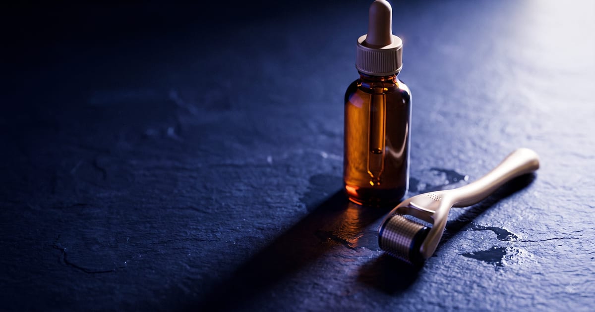 An amber pharmacy dropper bottle on wet slate casting a long shadow, beside a champagne-gold titanium derma roller